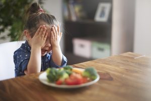 Criança com autismo cobrindo o rosto, recusando comer vegetais em um prato.