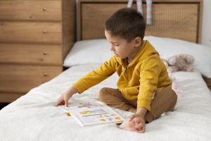 Menino pequeno sentado na cama lendo um livro ilustrado em seu quarto.