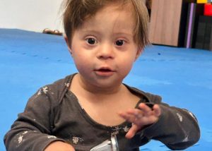 Bebê pequeno com traços de síndrome de Down sentado no chão, olhando para a câmera e segurando um objeto.