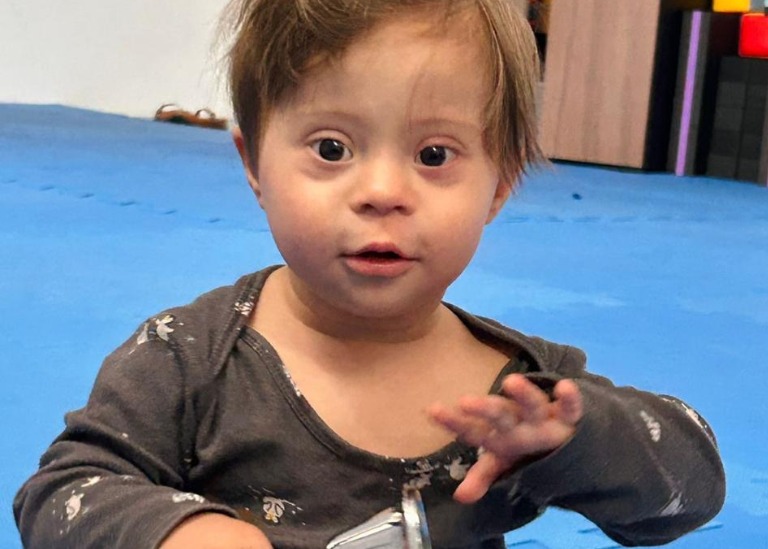 Bebê pequeno com traços de síndrome de Down sentado no chão, olhando para a câmera e segurando um objeto.